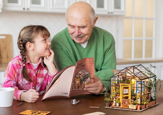 A grandpa wearing green cardigan helping his granddaughter wearing a pink flannel with her DIY miniature house kit.