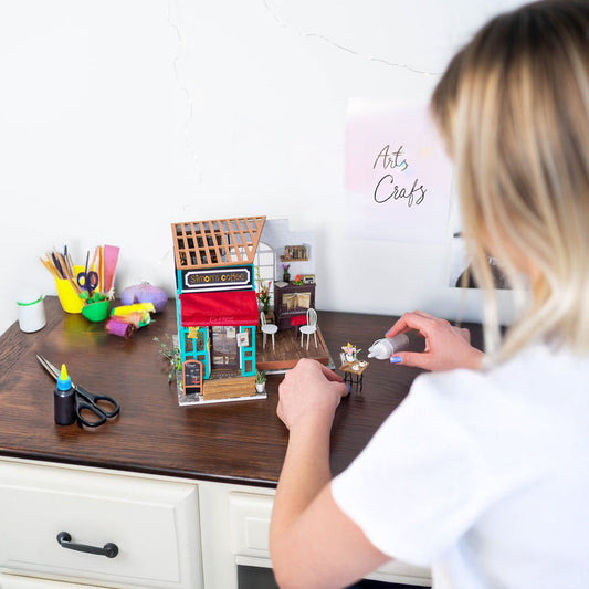 A woman putting crafts glue on a miniature furniture. She is wearing white shirt and has blond hair. There is arts and crafts supplies on her working desk.