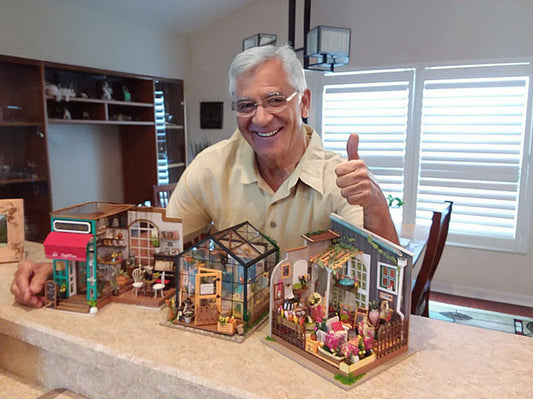 A handsome elderly man showing off his collections of miniature DIY house kit. He has gray hair, glasses, and wearing a light yellow polo shirt. He is smiling and seems very proud, throwing a big thumbs up.